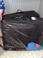 Two black storage bags containing fold out foam beds, placed on the floor next to a wall and an American flag.