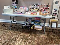 Full view of a gray plastic folding table with various craft items on top and board games underneath. The table is on a patterned rug in a craft room environment.
