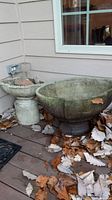 Two concrete fountain pieces, large oval basin on a pedestal base with a smaller vase-like piece beside it, on outdoor deck covered in leaves.