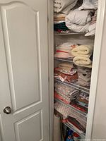Photo of a partially open closet door revealing shelves filled with various folded bed linens, towels, tablecloths, and cleaning supplies on the bottom shelf in a bathroom closet.