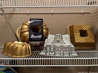 Shelf view showing four bundt pans: three gold Nordic Ware 75th Anniversary pans with fluted and geometric designs plus one silver gingerbread house shaped pan