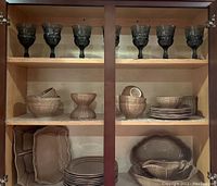 Cabinet showing upper shelf with seven blue goblets and middle shelves with brown glazed stoneware bowls and plates, lower shelves with platters and casserole dishes.