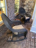 Pair of black rattan rocking chairs with beige upholstered cushions on front porch with brick floor and stone wall background.