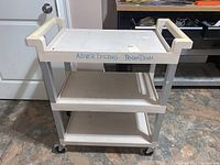 Front view of the Rubbermaid Tuffmade white rolling cart showing all three shelves, handles, and caster wheels.