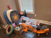Overview of toys showing boogie boards, pool noodle, frisbee, sports balls, and boxed toy supplies next to the Junior Road Builder excavator ride-on toy