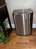 Side view of the chrome motion sensor trashcan placed on a wooden floor next to furniture and plants.