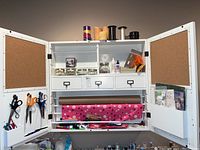 Open white wooden wall cabinet showing three small drawers, shelves with craft supplies including candles, jars with beads, glues, ribbons, and scissors attached to interior doors with pen holder and rolls of wrapping paper below.