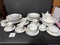 Photo showing full set including plates stacked at the back, teacups and saucers arranged in front, teapot, sugar bowl, cream pitcher, and serving bowl with lids all displayed on black surface.
