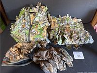 Ghillie suit jacket, gloves, and hat displayed on a black surface showing leaf camouflage pattern.