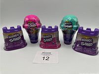 Five containers of Kinetic Sand including purple sandcastle containers and ice cream cone shaped containers shown in packaging.