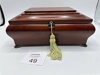 Closed wooden music jewelry box with lock and decorative cream tassel, showing wood finish and brass feet.