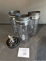 Three transparent plastic Ninja blending cups with black lids and one stainless steel blade attachment with black base, arranged on dark surface.