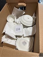 Box containing white ceramic plates and teacups with pink flower and green stem designs. Items are wrapped in paper towels for protection.