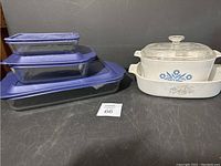 Five glass bakeware pieces including three blue-lidded Pyrex glass dishes and two white Corning Ware dishes with floral designs