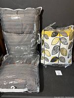 Two throw pillows in plastic packaging, one gray checkered with buttons, one white with yellow and black leaves.