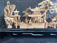 Front view of a detailed wood carving village including pagodas on elevated terrain, trees, and small figures, all inside a display case.