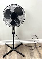 Full view of the oscillating standing fan showing black blades, protective grill, adjustable stand, and power cord on wooden floor against white wall.