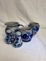 Set of five blue pottery items including one bowl, two pouring jugs, and salt and pepper shakers arranged on a white textured background.