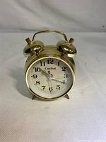Front view of brass Cardinal alarm clock showing white dial with black Arabic numbers and twin bells on top.