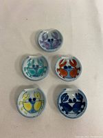 Top view showing five circular ceramic chopstick rests with handpainted crab faces in different colors: light purple, light blue, orange-red, yellow and dark blue.