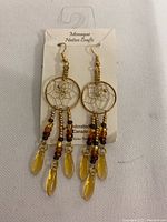 Front side photo of dream catcher earrings on branded card, showing circular metal hoop with webbing and dangling beaded strands with yellow feather charms.