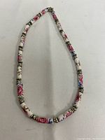 Full view of white necklace with hand painted pink rose porcelain beads and metal spacers, arranged in an oval shape on white fabric.