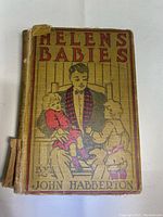 Front cover of the book showing the title 'Helen's Babies' and author John Habberton, with an illustrated image of a man holding two children. Cover shows heavy wear and tear, particularly along the spine and edges.
