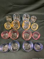 Top down view of all 16 glasses in four color groups (clear, yellow, pink/red, purple/blue) arranged in pairs of small and large glasses, laid on a dark background.