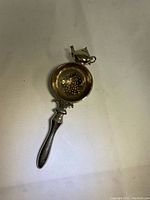 Silver-plated over tea cup tea strainer lying flat, showing perforated bowl and decorative teapot on top.