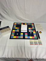 Photo showing the game board with a stack of blank sketch paper in the center, three black pencils, a white numbered die and colorful category dice, a small sand timer, and printed rules sheet to the side. Items are arranged on a white surface.