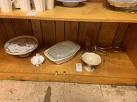 Wide view showing a porcelain cake stand, Pyrex casserole with lid, bowls, glass pitcher, and decorative wire holder on a wooden shelf.
