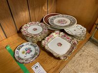 Assorted small dessert plates and square dishes with floral motif and gold rims on wooden shelf