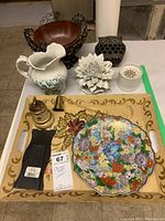 Overview of the lot items arranged on a table, showing the ceramic jug, white flower decor, painted tray, cloisonné plate, metal bowl, brass items, and the small white container.