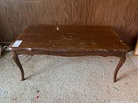 Front view of wooden coffee table showing worn top surface with scratches and stains, curved legs and scalloped edges.