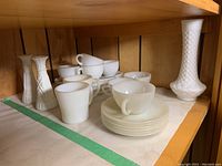 Overview of two small white milk glass bud vases, multiple Fire King cups, saucers, and white luncheon plates on shelf