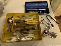 Overall view of flatware in plastic tray with forks, knives, and spoons, plus carving set box and serving utensils arranged on table