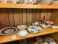 Shelf displaying various porcelain and china serving dishes, platters, and a teapot with floral designs and gold accents