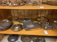 Wide view of multiple silverplate trays and covered serving dishes on wooden shelves, showing tarnish and varying designs.
