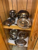 Wide shot of silverplate serving ware on wooden shelves including trays, pitcher, covered chafing dish, ice bucket, and silent butler tray showing tarnish.