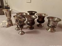 Frontal view of all pieces arranged on surface: jigger at left, two small goblets in front, five pedestal cups behind.