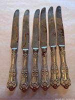 Full view of seven knives with ornate Birks sterling silver handles and rusted stainless steel blades arranged in a row.