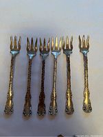 Six ornate sterling silver cocktail forks with monogram and three tines, arranged side by side