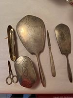 The full vanity set laid out on a flat surface showing hand mirror, hairbrush, nail file, covered item missing brush, rusted scissors, broken comb, filing box, and tray/case all with engraved monogram patterns.