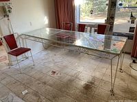 View of large vintage metal and glass dining table with four red vinyl and metal frame chairs arranged around it. Visible wear includes rust and chipped paint on metal frame, and stain on one chair.