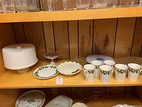 Shelf display of the lot showing six Royal Grafton mugs with holly design, a white ribbed covered cake stand, clear glass cake stands and various glass platters.