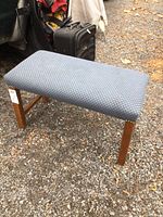 Rectangular upholstered bench standing on gravel ground with grey-blue fabric and wooden legs.