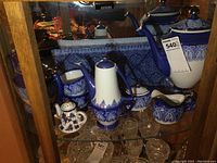 Front view of the porcelain tea set on a glass cabinet shelf with blue and white pattern details and crystal glassware below.