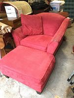 Red oversized armchair with cushion and matching ottoman shown from angled side view in a cluttered space.