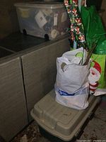 Photo showing two clear plastic totes stacked on a cabinet, a white plastic bag with twig-like decor, and a green bag with Santa print, indicating storage of holiday crafts and decorations.
