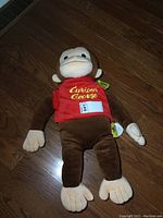 Curious George plush monkey lying on wooden floor showing front view with face and embroidered red shirt visible.